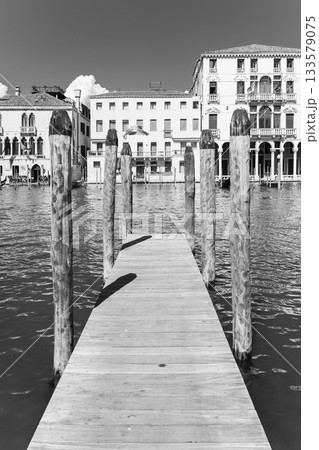 A wooden jetty extends into the calm waters of Venice, surrounded by historic buildings. The sun casts shadows while fluffy clouds float above the serene landscape, inviting exploration. A wooden jetty extends into the calm waters of Venice, surrounded by historic buildings. The sun casts shadows while fluffy clouds float above the serene landscape, inviting exploration. 133579075