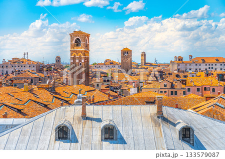 This aerial view captures the intricate rooftops of Venice, showcasing its historic architecture, bell towers, and a vibrant cityscape under a bright blue sky with scattered clouds. 133579087