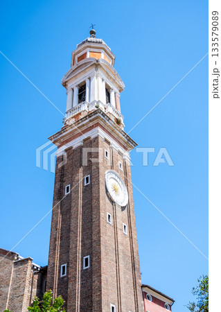 The Campanile dei Santi Apostoli rises 47 meters into the Venetian sky, showcasing its brick façade and prominent clock against a bright blue backdrop in the Cannaregio district. The Campanile dei Santi Apostoli rises 47 meters into the Venetian sky, showcasing its brick façade and prominent clock against a bright blue backdrop in the Cannaregio district. 133579089