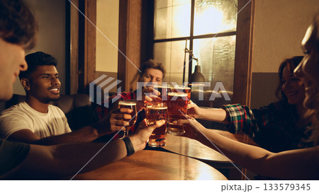 Friends raising beer glasses together at pub booth in warm scene 133579345