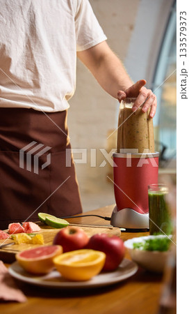 Man blending smoothie in compact blender with fresh fruit and greens on table. 133579373