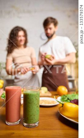 Pink and green smoothies standing on table while couple prepares fruit nearby Pink and green smoothies standing on table while couple prepares fruit nearby 133579374