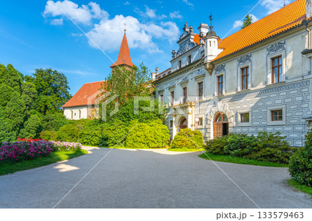 Pruhonice Castle showcases the stunning Church of the Nativity of the Virgin Mary, surrounded by vibrant greenery and colorful flowers under a clear blue sky. Visitors admire the architecture. 133579463