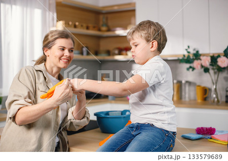 Blonde boy and adult woman cleaning in a kitchen and wearing orange gloves. Caucasian boy helping his mother to do housework. Mother helping her son to wear rubber gloves. 133579689