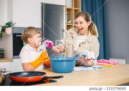 Blonde boy and adult woman cleaning in a kitchen and wearing orange gloves. Caucasian boy helping his mother to do housework. Blue busket with different rugs and detergents on a table. 133579690