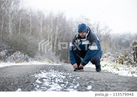Adult sporty man at walk in winter clothing tying shoelaces and snowy road at park. Cold weather and snowy season. Outdoors. Adult sporty man at walk in winter clothing tying shoelaces and snowy road at park. Cold weather and snowy season. Outdoors. 133579948