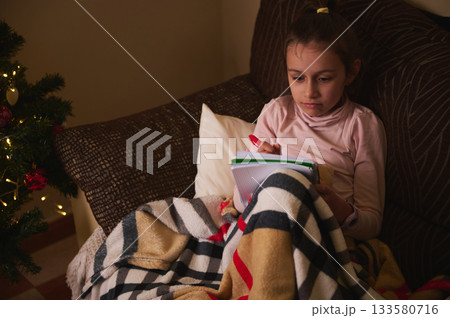Child Writing in Notebook on Couch With Blanket Near Christmas Tree Lights 133580716