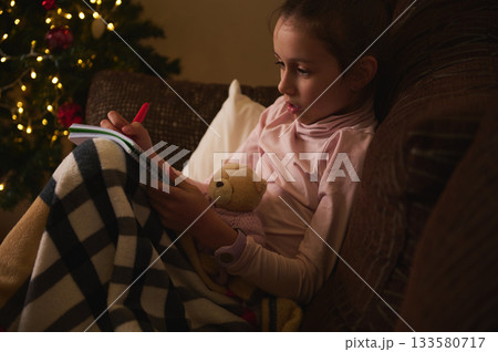 Child Reading Hobby By Christmas Tree On Sofa With Teddy Bear And Cozy Blanket 133580717