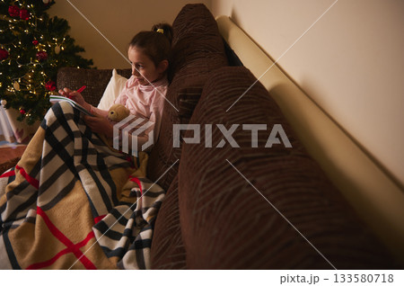 Cozy Christmas Night: Girl Reading on Couch with Blanket and Teddy Bear Near Tree Cozy Christmas Night: Girl Reading on Couch with Blanket and Teddy Bear Near Tree 133580718