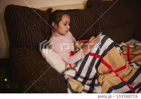 Cozy Girl Writes in Notebook While Snug Under Blanket on Couch Cozy Girl Writes in Notebook While Snug Under Blanket on Couch 133580719