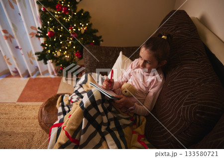 Cozy Child Writing Notebook on Couch With Teddy Near Christmas Tree 133580721