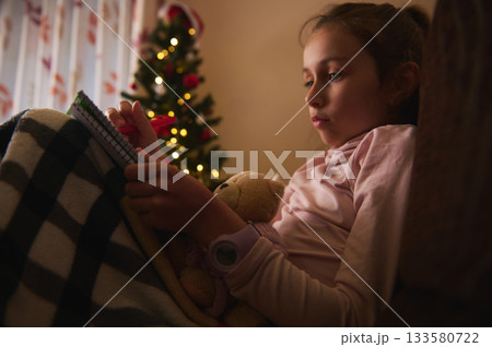 Cozy Christmas Night: Child Reading Notebook With Teddy Bear By a Decorated Christmas Tree 133580722