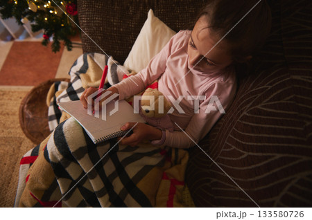 Little Girl Writing in Notebook on Sofa with Teddy Bear and Cozy Blanket by Christmas Tree Little Girl Writing in Notebook on Sofa with Teddy Bear and Cozy Blanket by Christmas Tree 133580726