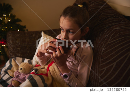 Young Girl Relaxing On Couch With Wooden Toy Instrument Near Christmas Tree 133580733