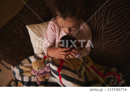 Young Child Sits Cozy On Couch Holding Wooden Cup, Warm Blanket And Plush Bear Nearby 133580734