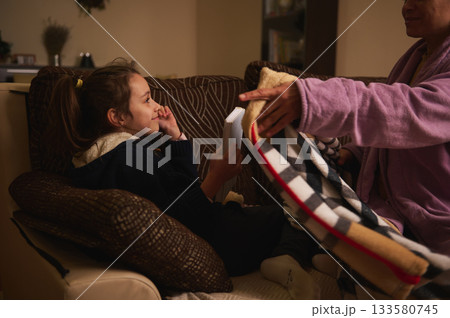 Young Girl Reading a Book on Sofa as Grandparent Covers Her with a Blanket in Cozy Living Room 133580745