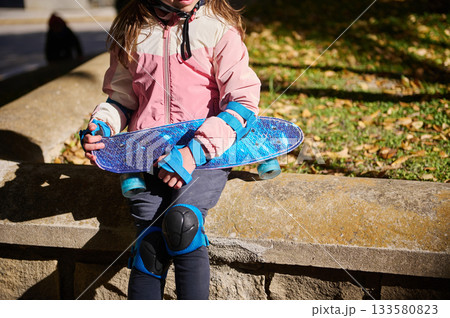 Kid With Blue Skateboard On Park Wall In Fall Light Wearing Pink Jacket And Knee Pads Kid With Blue Skateboard On Park Wall In Fall Light Wearing Pink Jacket And Knee Pads 133580823