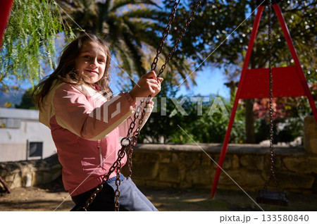 Joyful Girl On A Swing Outdoors, Sunlit Park Scene With Colorful Playground Equipment 133580840
