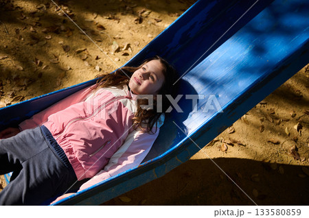 Girl Relaxing On Blue Playground Slide In Pink Jacket Outdoors 133580859