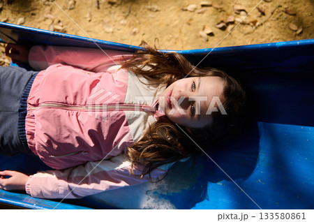 Playful Girl Lying in a Blue Slide Outdoors in Pink Jacket on a Sunny Day Playful Girl Lying in a Blue Slide Outdoors in Pink Jacket on a Sunny Day 133580861