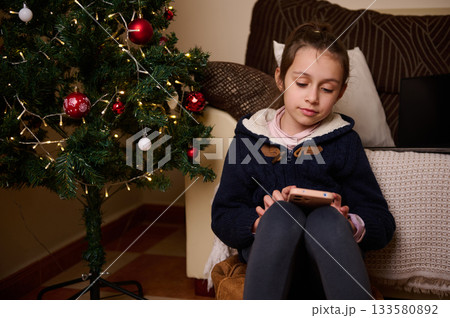 Girl By Christmas Tree Uses Phone In Cozy Living Room During Holiday Season Girl By Christmas Tree Uses Phone In Cozy Living Room During Holiday Season 133580892
