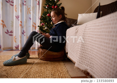 Young Girl Relaxed By Christmas Tree, Sitting on Pouf with Tablet in Cozy Living Room Young Girl Relaxed By Christmas Tree, Sitting on Pouf with Tablet in Cozy Living Room 133580893