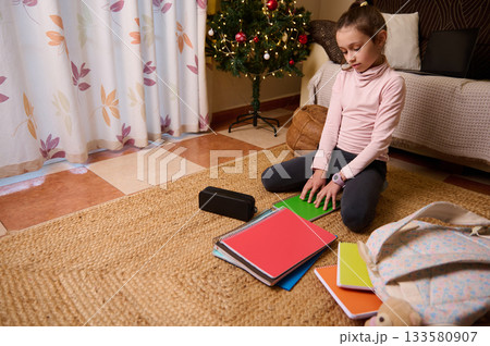 Young Girl Studying On Rug With Colorful Notebooks Near Christmas Tree Young Girl Studying On Rug With Colorful Notebooks Near Christmas Tree 133580907