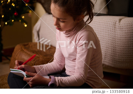 Girl Writing in Notebook by Christmas Tree Lights in Cozy Room 133580918