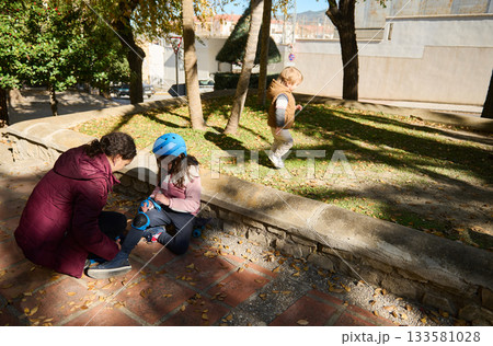 Family Helps Little Girl Tie Skates In Park While One Child Walks Nearby On Sunny Day 133581028