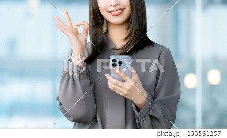 スマートフォンを持ってOKポーズをする笑顔の女性・ビジネスウーマン 133581257