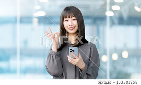 スマートフォンを持ってOKポーズをする笑顔の女性・ビジネスウーマン 133581258
