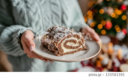 Woman in cozy sweater holds a slice of buche de noel on a plate, decorated Christmas tree softly blurred in the background, creating a festive atmosphere 133581391