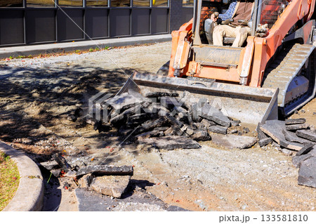 Machinery skid steer breaks apart asphalt in parking lot as worker oversees removal process 133581810