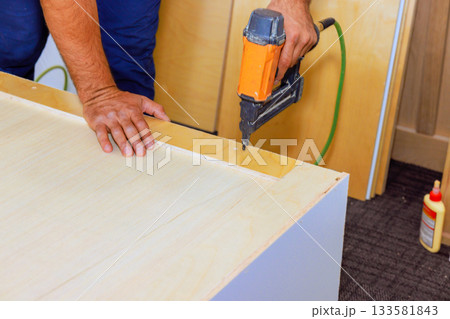 Carpenter uses nail gun on piece of wood, working on furniture assembly Carpenter uses nail gun on piece of wood, working on furniture assembly 133581843