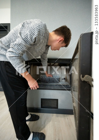 Young man carefully cleaning the inside of a sleek fireplace in a modern living room, focusing on removing ash and debris for a tidy appearance Young man carefully cleaning the inside of a sleek fireplace in a modern living room, focusing on removing ash and debris for a tidy appearance 133583963