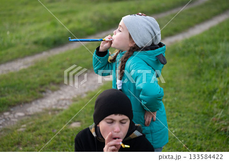 Two children enjoy a playful moment outdoors, blowing whistles and creating joyful sounds on a grassy path during the golden hour of late afternoon Two children enjoy a playful moment outdoors, blowing whistles and creating joyful sounds on a grassy path during the golden hour of late afternoon 133584422