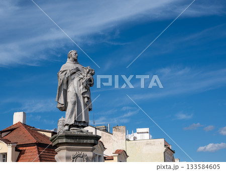 Prague, czech republic, august 9, 2023. Stone statue of a saint wearing robes and holding flowers, standing on a pedestal with an ornate base against a bright blue sky 133584605