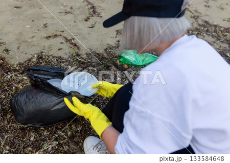 Volunteer collecting plastic waste and litter into a black trash bag during beach cleanup, highlighting pollution, environmental protection, sustainability, and eco awareness. Volunteer collecting plastic waste and litter into a black trash bag during beach cleanup, highlighting pollution, environmental protection, sustainability, and eco awareness. 133584648