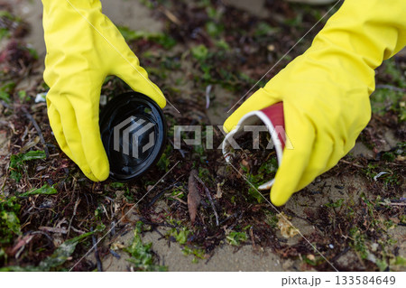 Hands in yellow gloves collecting plastic cup waste during beach cleanup, highlighting pollution, environmental protection and ocean sustainability. Hands in yellow gloves collecting plastic cup waste during beach cleanup, highlighting pollution, environmental protection and ocean sustainability. 133584649
