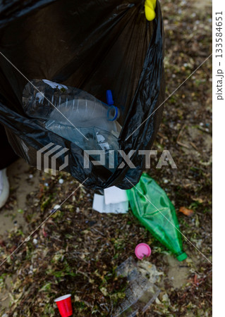 Volunteer collecting plastic waste and litter into a black trash bag during beach cleanup, highlighting pollution, environmental protection, sustainability, and eco awareness. 133584651
