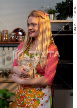 A smiling woman in a floral apron holds fresh ingredients in a modern kitchen, ready to cook A smiling woman in a floral apron holds fresh ingredients in a modern kitchen, ready to cook 133584866