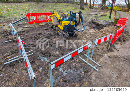 Mini excavator at fenced-off construction site in park during ground excavation work Mini excavator at fenced-off construction site in park during ground excavation work 133585629