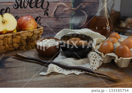 High-quality baking ingredients like fresh apples, cocoa powder, white flour, and brown eggs arranged with a dark wooden spoon highlighting home cooking and sweet food preparation 133585754