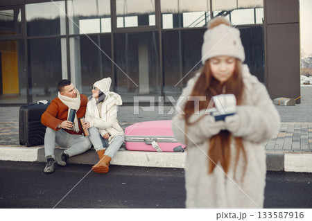 Little girl with her parents going on vacation. Portrait of a girl standing in front of photo and and her parents sitting behind. Family wearing warm clothes. Little girl with her parents going on vacation. Portrait of a girl standing in front of photo and and her parents sitting behind. Family wearing warm clothes. 133587916