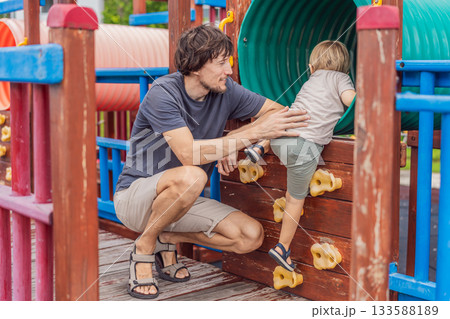 A man playing with his small son on a playground, laughing and engaging in active fun together in a bright outdoor setting. Family bonding, early childhood development and healthy outdoor lifestyle 133588189