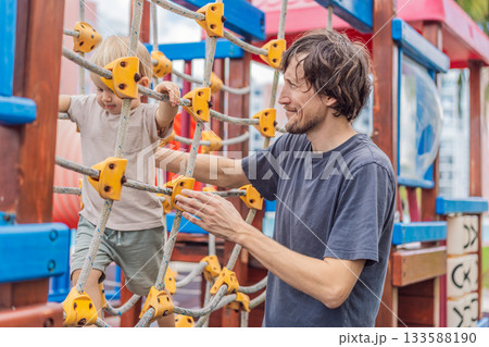A man playing with his small son on a playground, laughing and engaging in active fun together in a bright outdoor setting. Family bonding, early childhood development and healthy outdoor lifestyle 133588190
