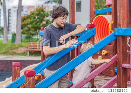 A man playing with his small son on a playground, laughing and engaging in active fun together in a bright outdoor setting. Family bonding, early childhood development and healthy outdoor lifestyle 133588192