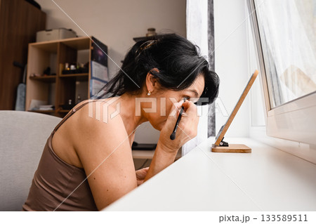 Side view of a mature Russian woman 50 years old sitting in a bedroom and applying eye makeup with a brush leaning toward a small mirror in soft calm light Side view of a mature Russian woman 50 years old sitting in a bedroom and applying eye makeup with a brush leaning toward a small mirror in soft calm light 133589511