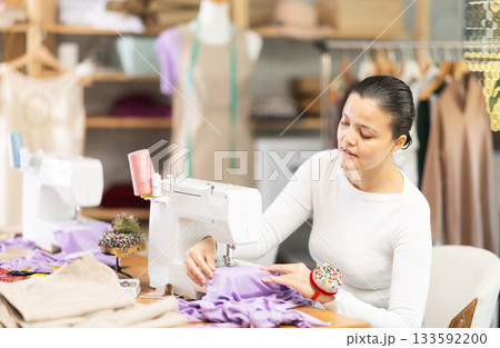 Professional young attentive woman seamstress sewing textile using sewing machine sitting at table at workplace in sewing studio Professional young attentive woman seamstress sewing textile using sewing machine sitting at table at workplace in sewing studio 133592200