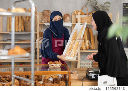 Muslim saleswoman gives baguettes to Muslim girl in bakery Muslim saleswoman gives baguettes to Muslim girl in bakery 133592548